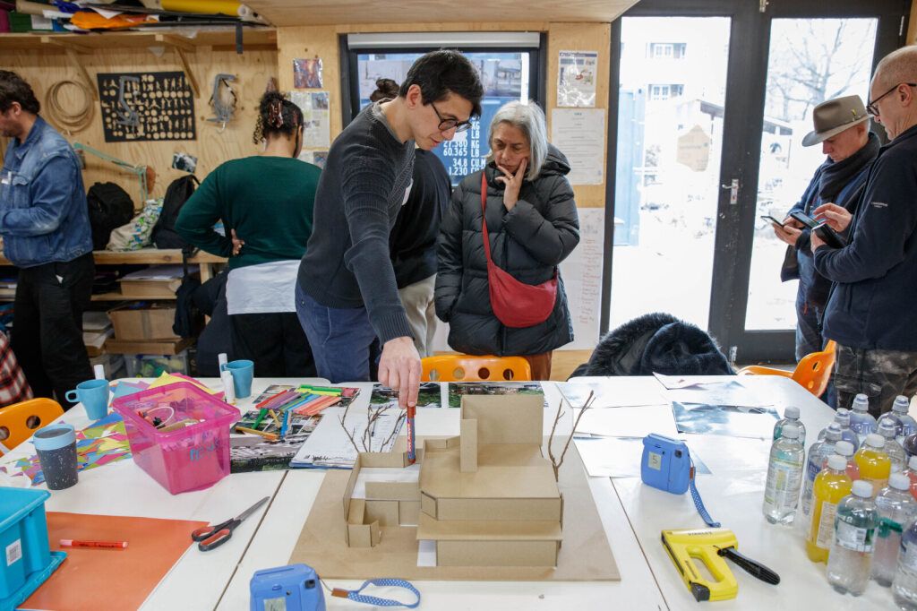 Two people stand over a table covered in pens and paper. One points to something with a pen while the other looks thoughtful.