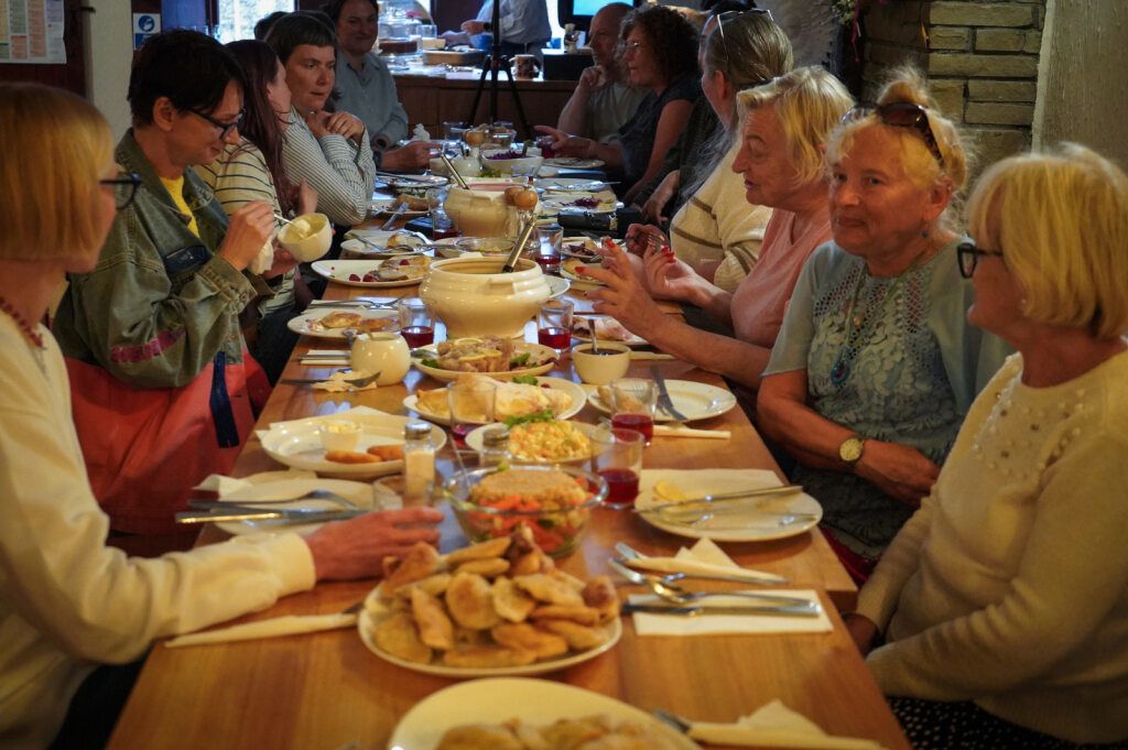 A shot taken from the head of a long dining table. 12 people sat either side talking and eating. Plates full of food run down the centre.