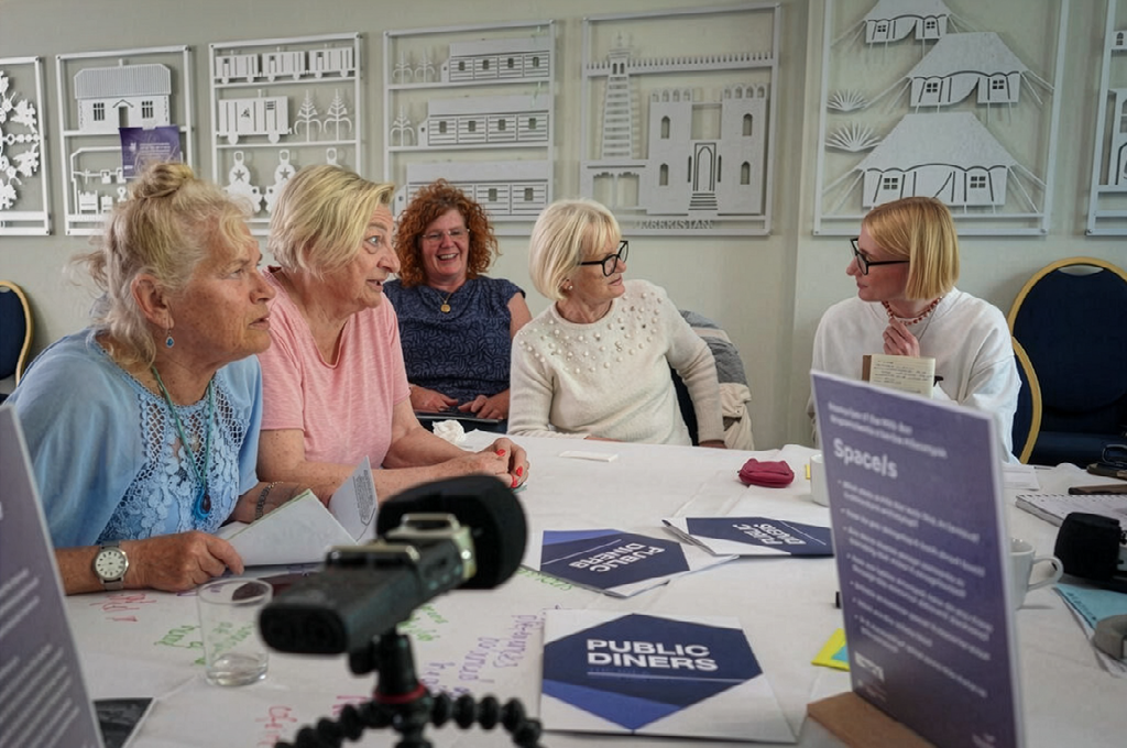 Five people sit round a table in conversation. Leaflets with the title Public Diners are spread on the table