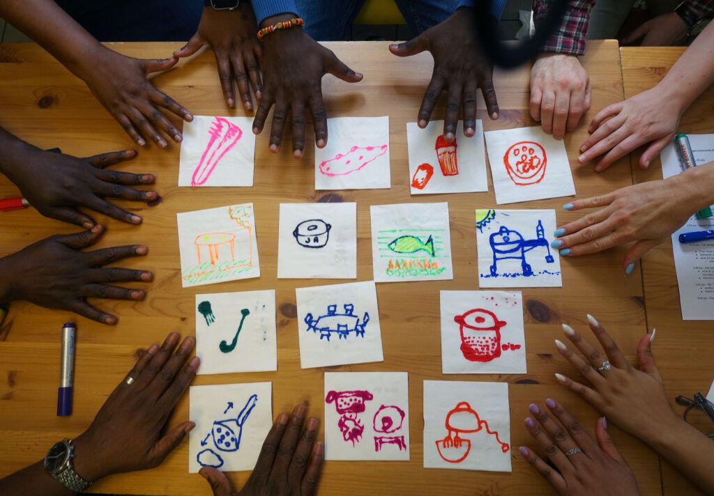 Fifteen square pieces of paper on a table, each with a hand drawn image of food or tables.