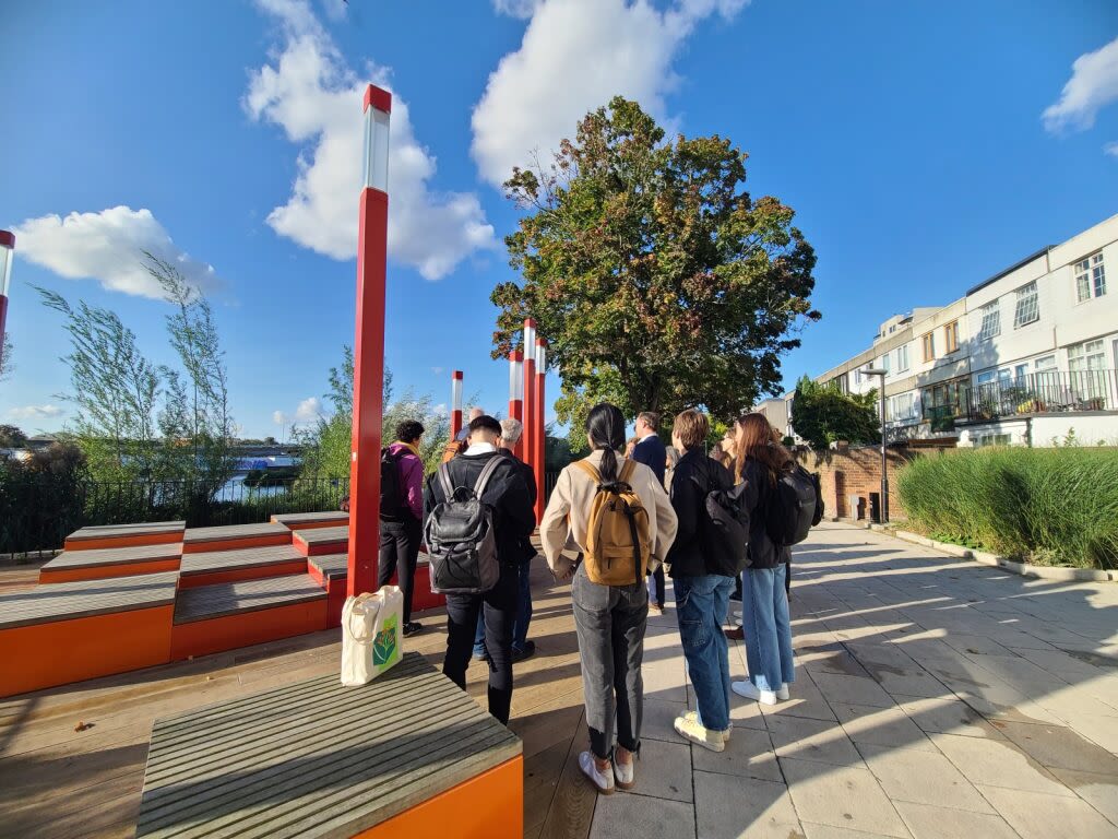 Group of people walking through a paved area in Thamesmead on a sunny day, with a tree and blue sky in the background.