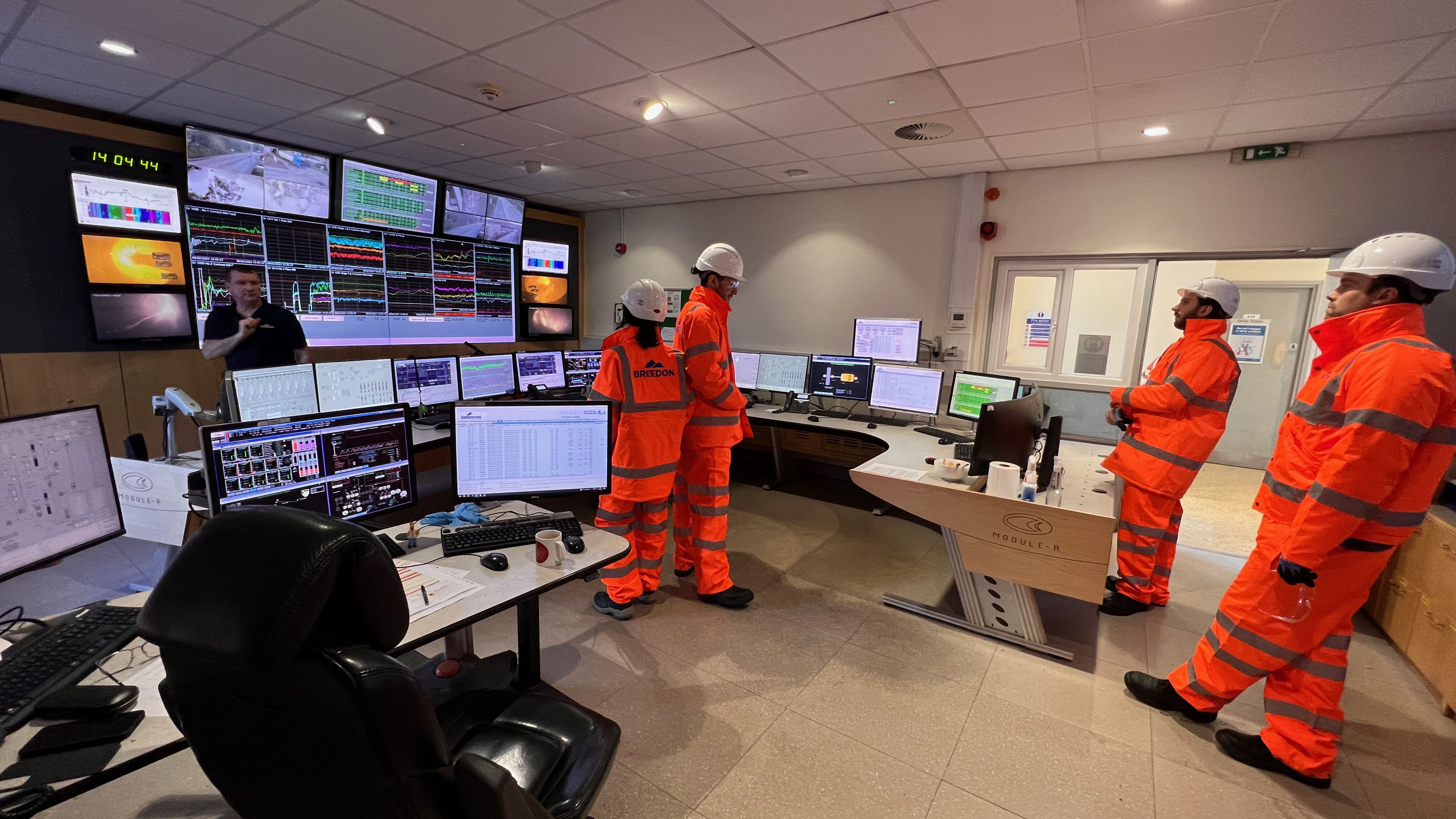 Workmen in hard hats standing in office with a large bank of data monitor screens